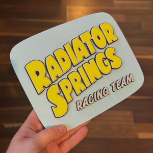 Puede incluir: Un letrero azul claro con las palabras "RADIATOR SPRINGS" en letras amarillas grandes con contornos negros. Debajo, el texto "RACING TEAM" est&aacute; en letras grises claras m&aacute;s peque&ntilde;as. El letrero es sostenido por una mano.