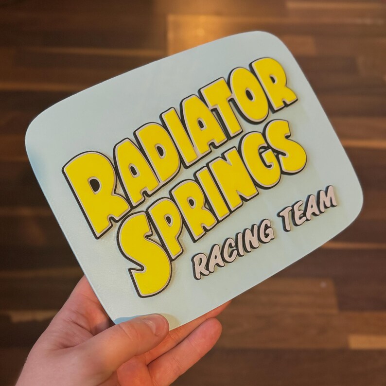 Puede incluir: Un letrero azul claro con las palabras "RADIATOR SPRINGS" en letras amarillas grandes con contornos negros. Debajo, el texto "RACING TEAM" est&aacute; en letras grises claras m&aacute;s peque&ntilde;as. El letrero es sostenido por una mano.