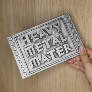 Peut inclure: Panneau rectangulaire argent&eacute; avec les mots "HEAVY METAL MATER" en relief. Le panneau a une bordure avec des rivets circulaires. Il est tenu par une main sur un fond en bois clair.
