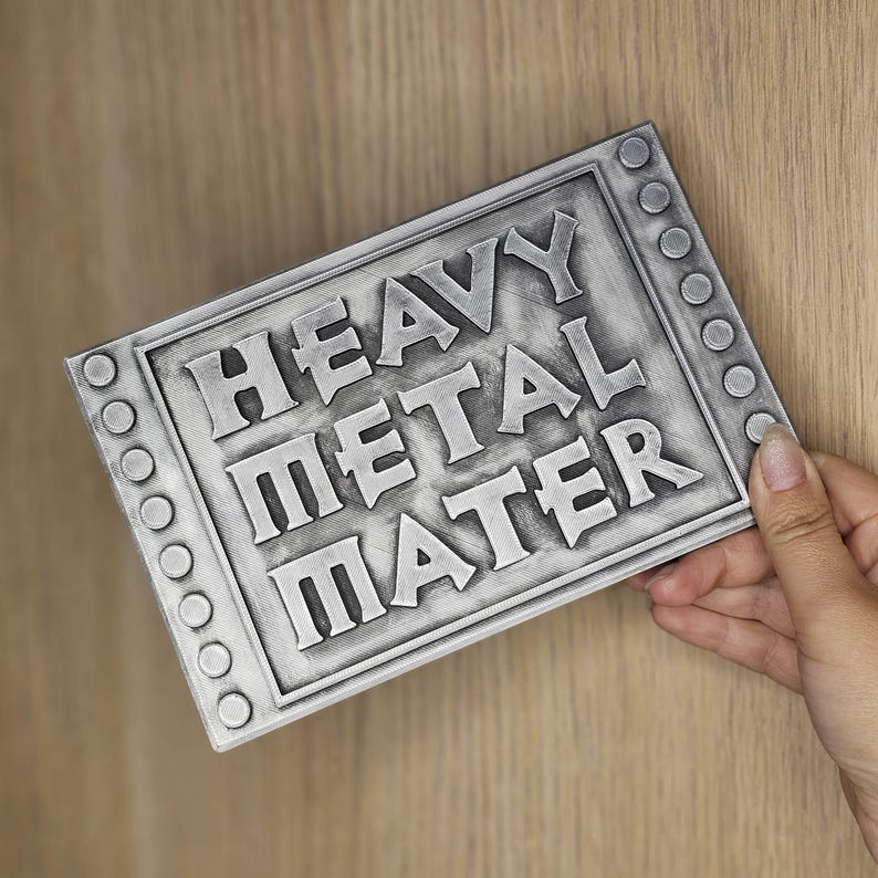 Peut inclure: Panneau rectangulaire argent&eacute; avec les mots "HEAVY METAL MATER" en relief. Le panneau a une bordure avec des rivets circulaires. Il est tenu par une main sur un fond en bois clair.
