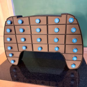 May include: A brown, 3D-printed advent calendar shaped like a video game controller. It features 24 numbered blue buttons and small drawers for each day of the countdown. The controller design includes a D-pad, buttons, and shoulder buttons.