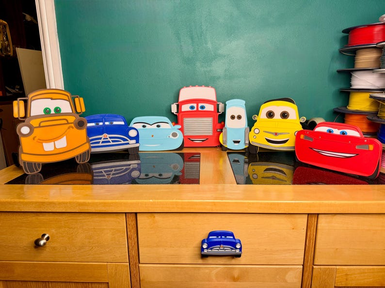 May include: A collection of seven colorful, flat, cartoon-style car-shaped wall decorations featuring various character designs, including a yellow tow truck, blue, red, and yellow vehicles, plus a small blue car-shaped drawer knob handle.
