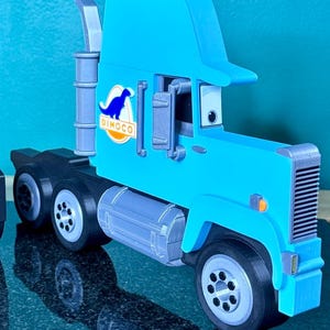May include: A light blue toy truck with the Dinoco logo on the side. The truck has black wheels, a silver exhaust pipe, and a detailed cab design. The truck is a 3D printed model.