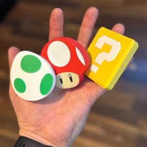 May include: A hand holding three colorful, cartoon-style objects: a red mushroom with white spots, a white egg with green spots, and a yellow block with a white question mark. The objects are likely from a video game.