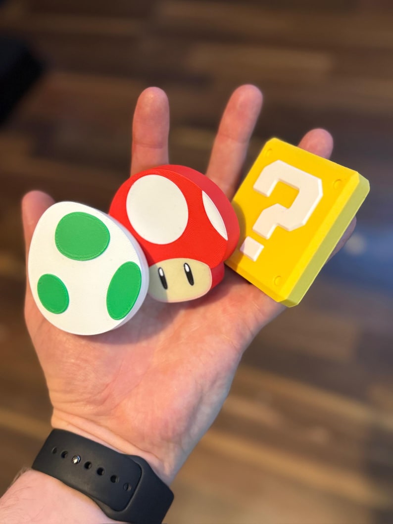 May include: A hand holding three colorful, cartoon-style objects: a red mushroom with white spots, a white egg with green spots, and a yellow block with a white question mark. The objects are likely from a video game.