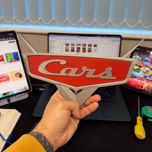 May include: A 3D-printed Cars logo sign, featuring a red background with the word "Cars" in silver lettering. The sign has a stylized wing design and is held in a person's hand. The background includes a laptop and other electronic devices.