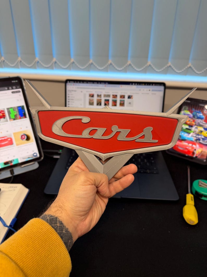 May include: A 3D-printed Cars logo sign, featuring a red background with the word "Cars" in silver lettering. The sign has a stylized wing design and is held in a person's hand. The background includes a laptop and other electronic devices.