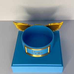 May include: A bright blue circular container mounted on a square blue base. The container features a gold-colored nameplate with the name Cain embossed in the center and a pair of gold-colored wings extending from the back of the rim.
