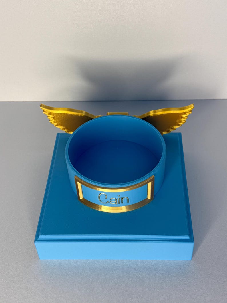 May include: A bright blue circular container mounted on a square blue base. The container features a gold-colored nameplate with the name Cain embossed in the center and a pair of gold-colored wings extending from the back of the rim.