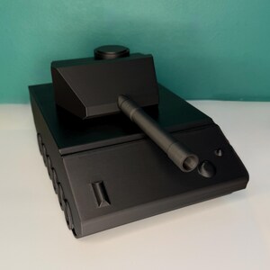 May include: A matte black toy tank model featuring a central turret with a long barrel, a flat hull, and visible wheel details along the side. The design is geometric and angular, with a smooth finish and a circular hatch on the turret top.