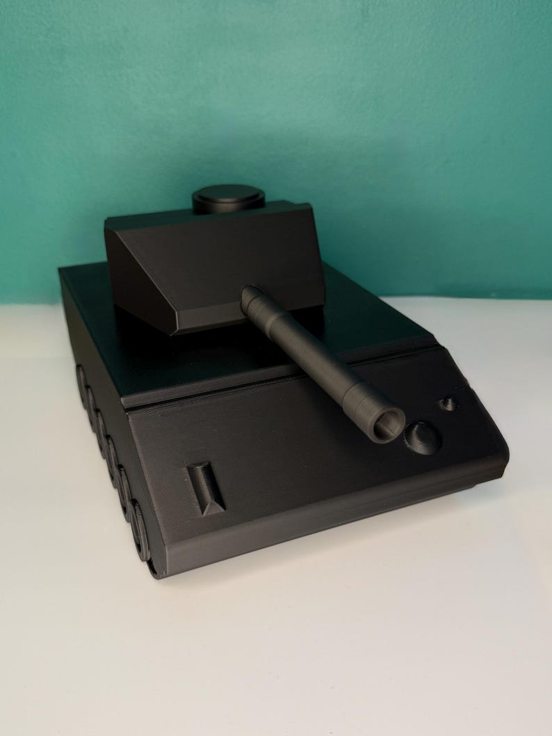May include: A matte black toy tank model featuring a central turret with a long barrel, a flat hull, and visible wheel details along the side. The design is geometric and angular, with a smooth finish and a circular hatch on the turret top.