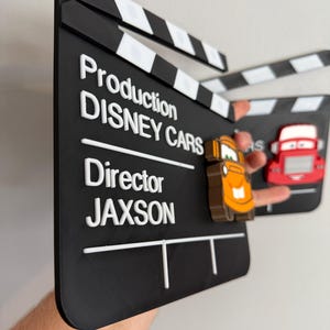 May include: A black clapperboard with white text that reads "Production DISNEY CARS Director JAXSON". The clapperboard has white and black stripes at the top and features 3D car characters. The clapperboard is held up.