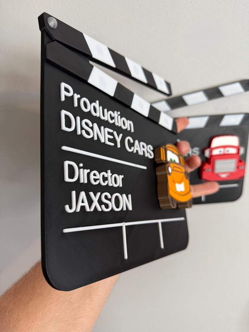 May include: A black clapperboard with white text that reads "Production DISNEY CARS Director JAXSON". The clapperboard has white and black stripes at the top and features 3D car characters. The clapperboard is held up.