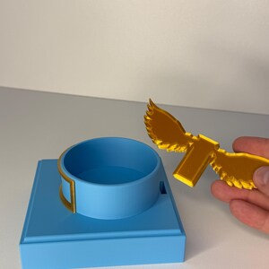 May include: A light blue square base with a circular ring holder and a separate gold-colored winged attachment. The base features a smooth matte finish, while the gold wing piece has a textured surface and a rectangular connector tab.