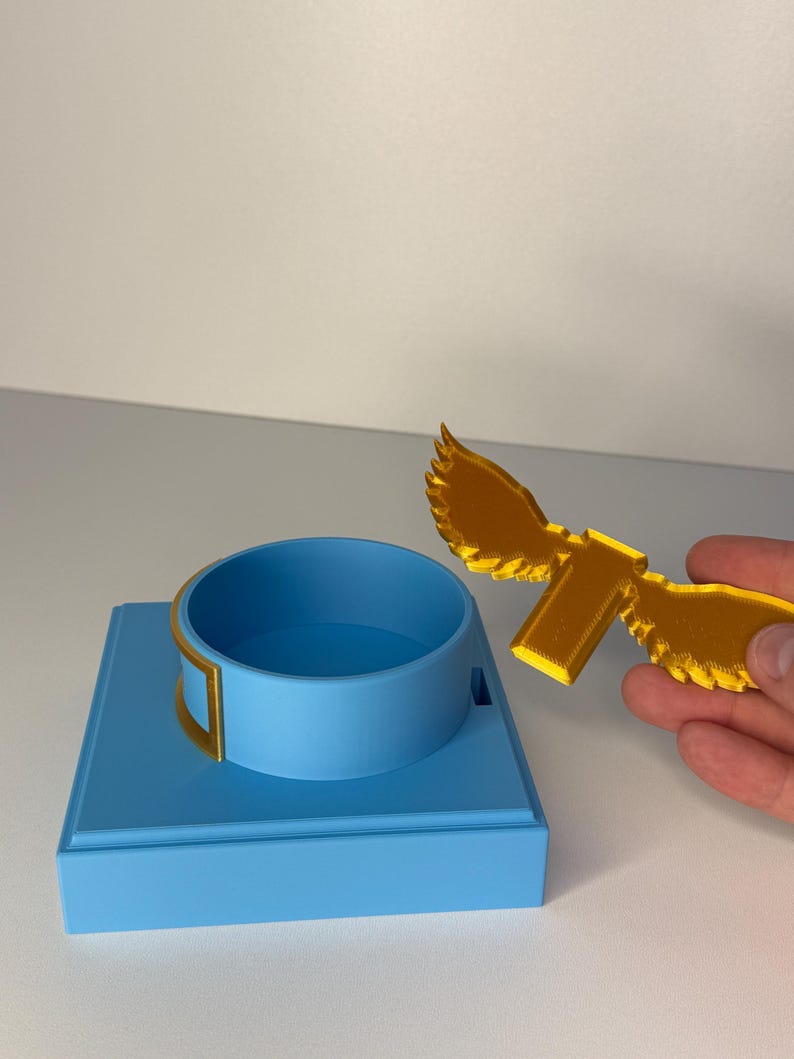 May include: A light blue square base with a circular ring holder and a separate gold-colored winged attachment. The base features a smooth matte finish, while the gold wing piece has a textured surface and a rectangular connector tab.