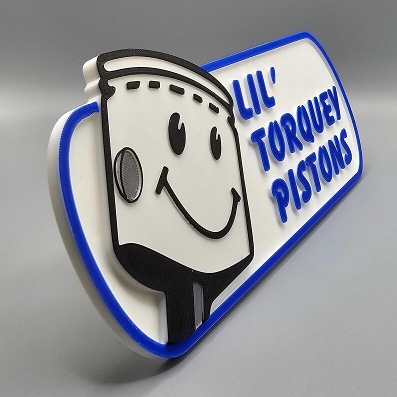 May include: A 3D-printed sign featuring a smiling cartoon character with the text "Lil' Torquey Pistons" in blue. The sign has a white background with a blue border and black accents, creating a playful and eye-catching design.
