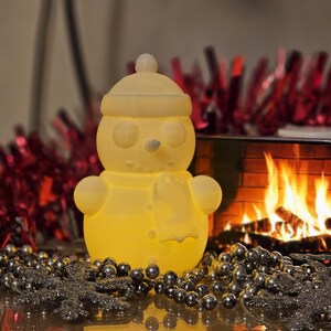 Yellow glowing 3D-printed snowman LED tealight holder, displayed on a festive table with red tinsel, silver beads, and a cosy fireplace in the background.