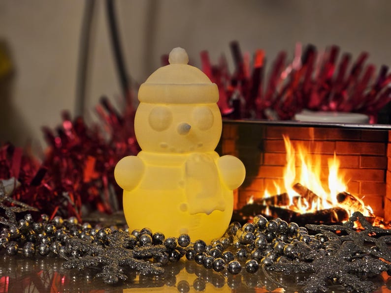 Yellow glowing 3D-printed snowman LED tealight holder, displayed on a festive table with red tinsel, silver beads, and a cosy fireplace in the background.