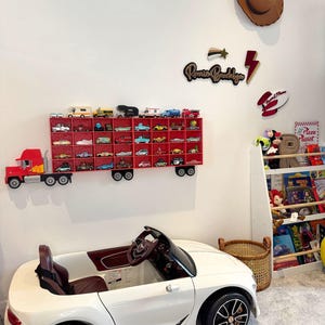 May include: A child's room featuring a white ride-on car with brown interior, a red truck-shaped display case filled with toy cars, and decorative wall art including a cowboy hat and a sign that says "Rome Brooklyn". A bookshelf is visible.