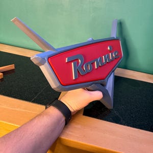May include: A 3D-printed sign with the name "Ronnie" in white script on a red background. The sign has a gray border and two gray fins extending from the top. The sign is held by a person.