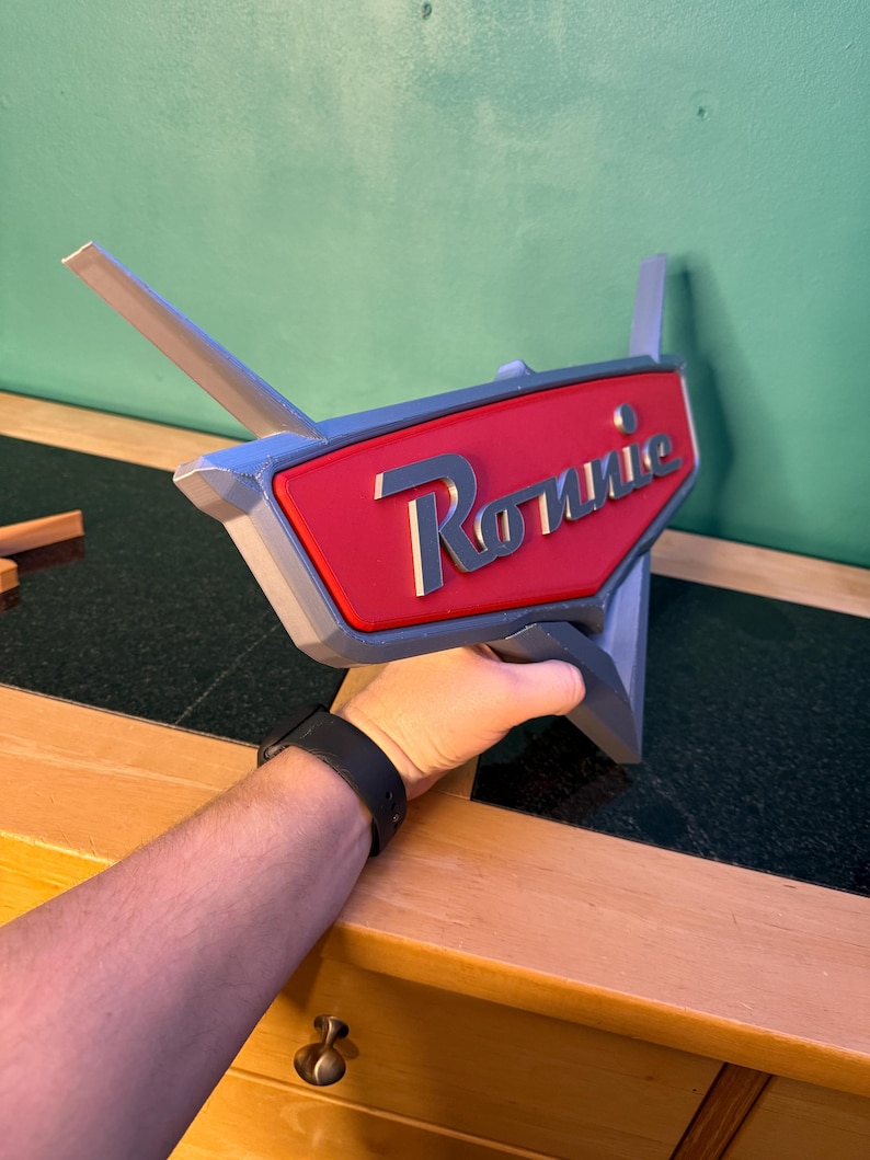 May include: A 3D-printed sign with the name "Ronnie" in white script on a red background. The sign has a gray border and two gray fins extending from the top. The sign is held by a person.