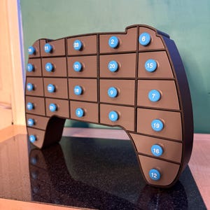 May include: A brown, game controller-shaped advent calendar with 24 numbered blue buttons. The controller has a black outline and is set against a green wall and a black surface.