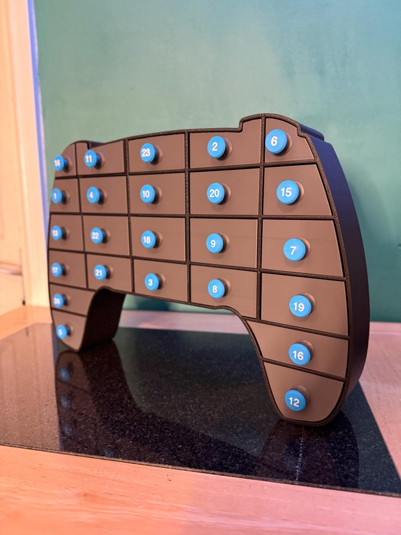 May include: A brown, game controller-shaped advent calendar with 24 numbered blue buttons. The controller has a black outline and is set against a green wall and a black surface.