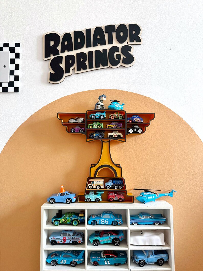 Puede incluir: Una exhibici&oacute;n de coches de juguete y un helic&oacute;ptero en una estanter&iacute;a blanca y una estanter&iacute;a decorativa. Las palabras "RADIATOR SPRINGS" est&aacute;n en letras negras. La pared est&aacute; pintada de color naranja claro.