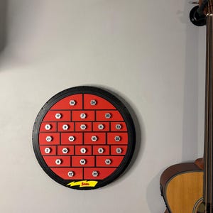 May include: A round, red and black advent calendar with 24 numbered compartments. The calendar is mounted on a gray wall, with a guitar visible on the right. The calendar has a black tire-like border and a yellow lightning bolt logo at the bottom.
