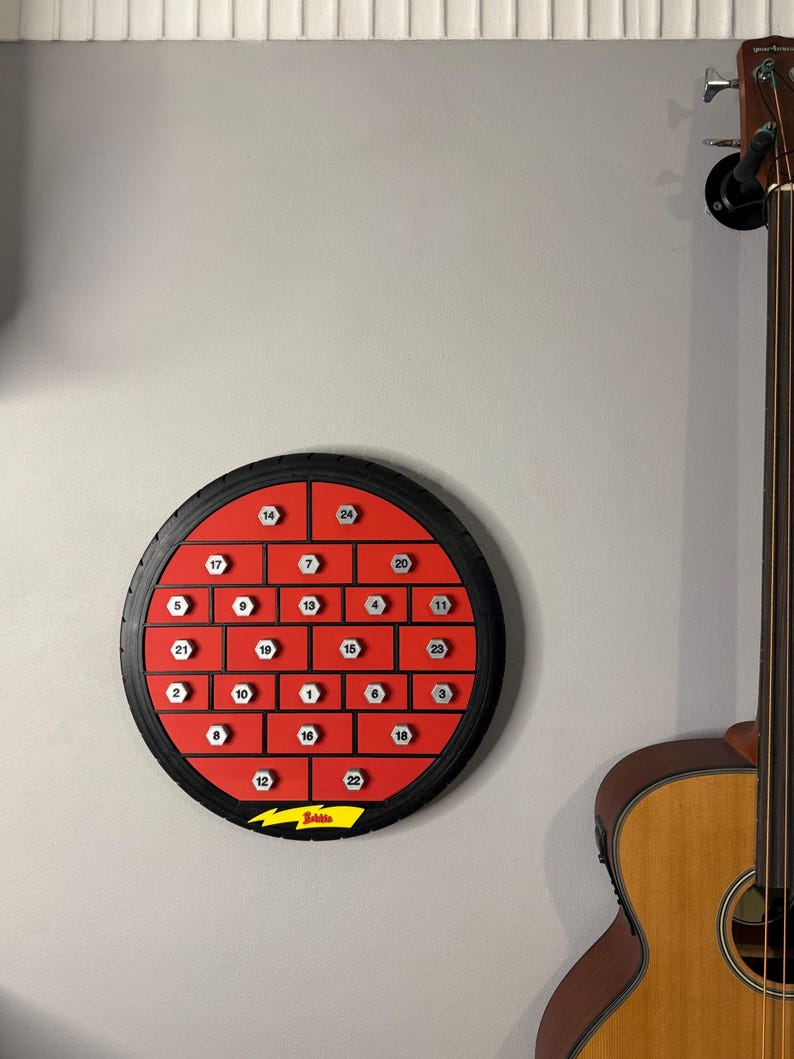 May include: A round, red and black advent calendar with 24 numbered compartments. The calendar is mounted on a gray wall, with a guitar visible on the right. The calendar has a black tire-like border and a yellow lightning bolt logo at the bottom.