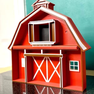 Puede incluir: Una granja roja en miniatura con adornos blancos y un techo marr&oacute;n. La granja presenta una gran abertura con una puerta marr&oacute;n y refuerzos cruzados blancos. Una peque&ntilde;a ventana es visible en el lateral. Es un objeto decorativo.