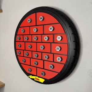 May include: A round, red advent calendar with a black tire-like frame. It features 24 numbered compartments with silver-colored, hexagonal knobs. The word "Battle" is visible on a yellow banner.