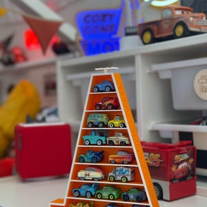 Op de afbeelding: Een oranje, driehoekige plank met een verzameling speelgoedauto's. De plank heeft meerdere niveaus, elk met een andere miniatuurauto. De auto's zijn in verschillende kleuren en ontwerpen, sommige met karakterdetails. De achtergrond bevat een neonreclame en andere speelgoedartikelen.