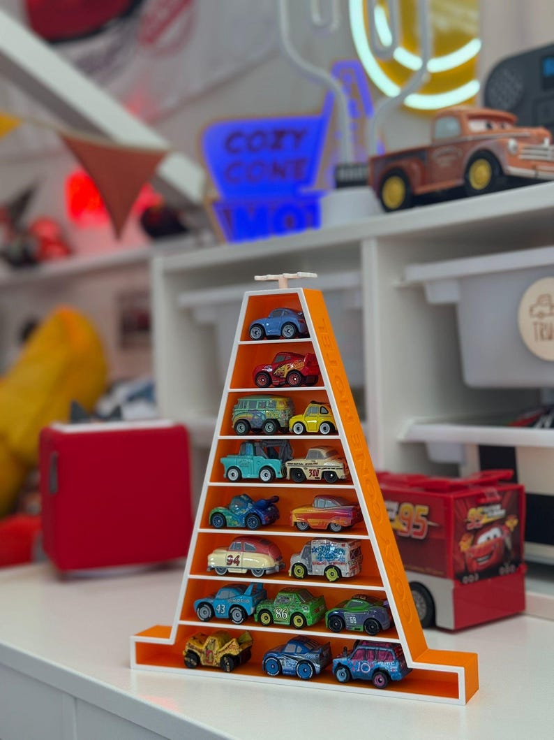 Op de afbeelding: Een oranje, driehoekige plank met een verzameling speelgoedauto's. De plank heeft meerdere niveaus, elk met een andere miniatuurauto. De auto's zijn in verschillende kleuren en ontwerpen, sommige met karakterdetails. De achtergrond bevat een neonreclame en andere speelgoedartikelen.