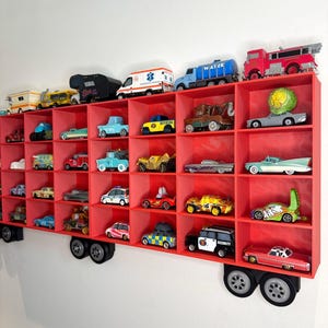 May include: A red wall-mounted shelf unit shaped like a truck, filled with toy cars of various colors and styles. The shelf has multiple cubby holes to display the toy cars. The toy cars include a fire truck, ambulance, and other vehicles.