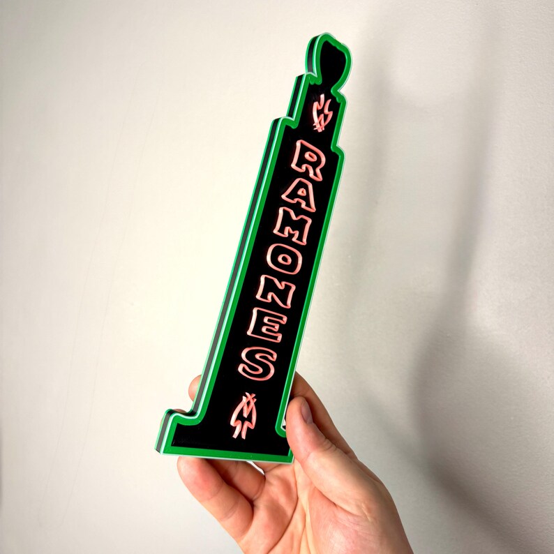 Puede incluir: Un letrero negro y verde con la palabra "RAMONES" en letras rosas. El letrero tiene un dise&ntilde;o de llama en la parte superior y un gr&aacute;fico de llama estilizado. El letrero est&aacute; sostenido por una mano.