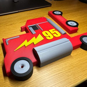 A 3D-printed red truck model inspired by Mack from Cars, featuring a yellow lightning bolt and the number 95. The parts are laid flat, showing two wheels, grey detailing, and black tires, all on a wooden surface.