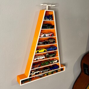 May include: An orange and white wall-mounted display shelf shaped like a traffic cone, filled with miniature toy cars. The shelf has multiple levels to hold the cars. The text "CONE MORE" is printed on the side of the shelf.