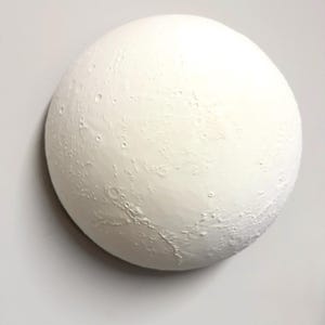 May include: A spherical wall sculpture featuring a textured, matte white surface that mimics the cratered topography of the moon. The object has a three-dimensional relief design with various circular indentations and raised ridges across its face.