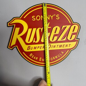 Puede incluir: Un letrero redondo rojo y amarillo con el texto "SONNY'S RUST-EZE BUMPER OINTMENT REAR END FORMULA". El letrero mide aproximadamente 94 cm de alto. Se sostiene una cinta m&eacute;trica amarilla contra el letrero.