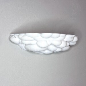 May include: A white, cloud-shaped wall shelf with a textured, bubbly surface. The shelf is mounted on a gray wall and features a small, circular emblem on the left side. This decorative shelf is designed to hold small items.