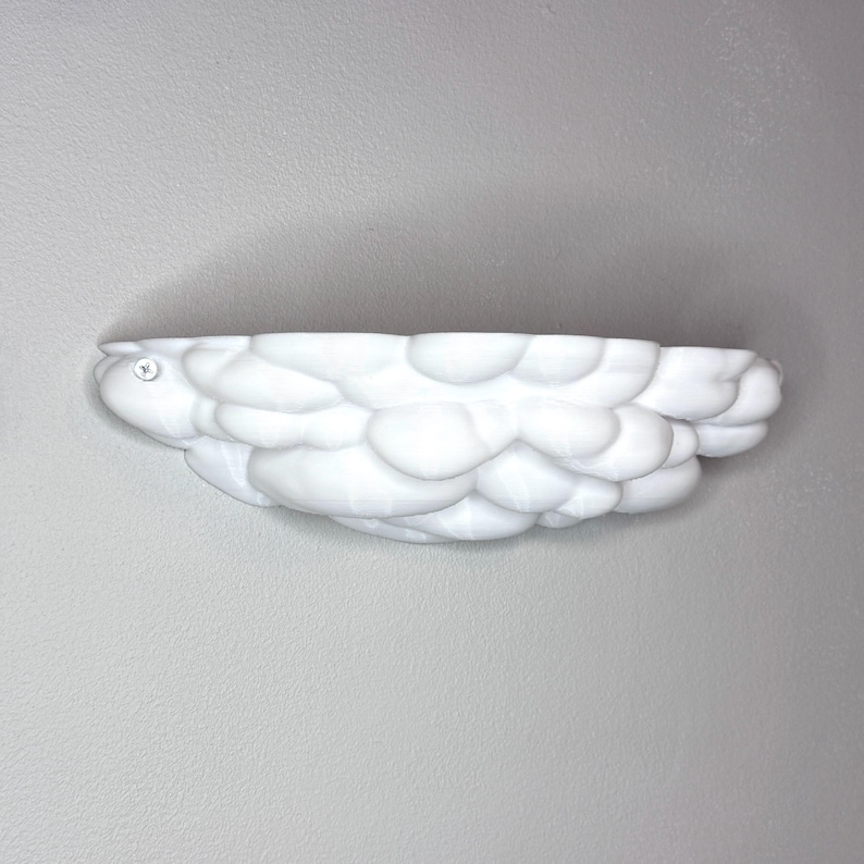 May include: A white, cloud-shaped wall shelf with a textured, bubbly surface. The shelf is mounted on a gray wall and features a small, circular emblem on the left side. This decorative shelf is designed to hold small items.