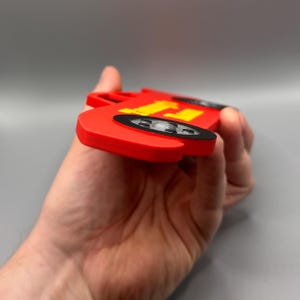 May include: A red toy car with yellow lettering and black wheels. The car is held in a hand, showcasing its side profile. The design is reminiscent of a classic race car.