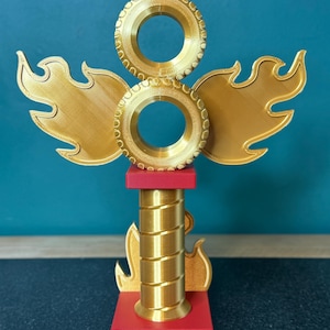 May include: A gold trophy with red detailing. It features two tyre-shaped elements, flame-like wings, and a central column. The base is inscribed with "WINNER" in gold. This trophy is likely for a racing or automotive competition, and is made of plastic.