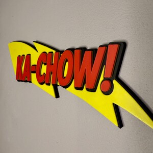 May include: A 3D-printed sign with the phrase "KA-CHOW!" in red, outlined in black, against a yellow lightning bolt shape. The sign is mounted on a gray wall.