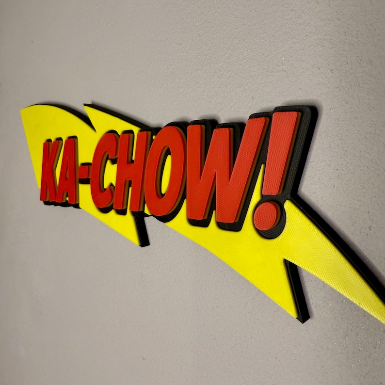 May include: A 3D-printed sign with the phrase "KA-CHOW!" in red, outlined in black, against a yellow lightning bolt shape. The sign is mounted on a gray wall.