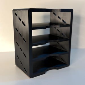 May include: A black, four-tiered shelf unit with a rectangular shape. The sides feature diagonal cut-out designs. The shelves are evenly spaced, and the unit has a curved bottom edge. The unit is made of a solid, matte black material.