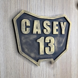 May include: A custom wall sign shaped like the state of Ohio featuring a dark charcoal background with a distressed gold border and raised gold lettering that reads CASEY 13. The sign has a textured finish and is mounted on a light wood surface.
