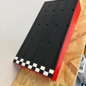 May include: A rectangular display stand with a black top and red sides. The top has multiple vertical grooves. The front edge features a checkered pattern of white and black squares. The stand is angled and appears to be made of wood.