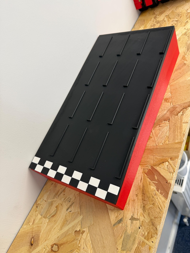 May include: A rectangular display stand with a black top and red sides. The top has multiple vertical grooves. The front edge features a checkered pattern of white and black squares. The stand is angled and appears to be made of wood.